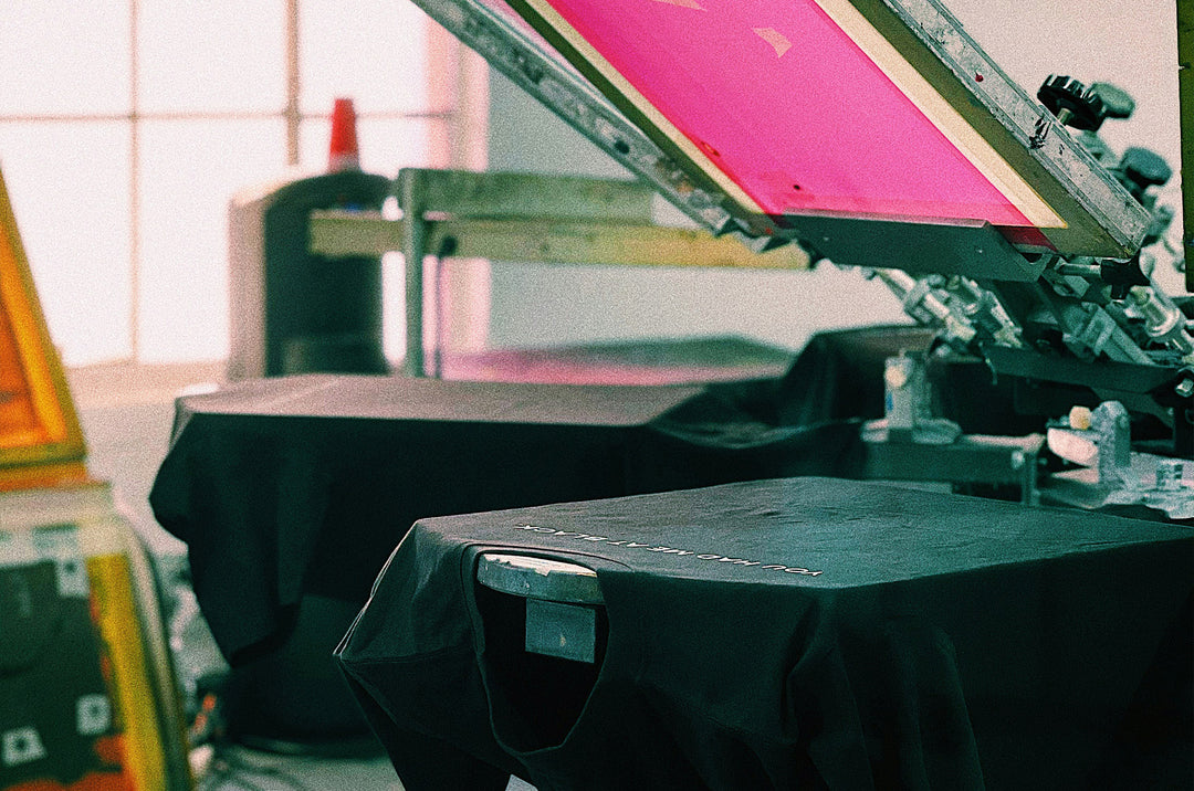 Screen printing press with a pink screen and black t-shirt in a workshop setting.